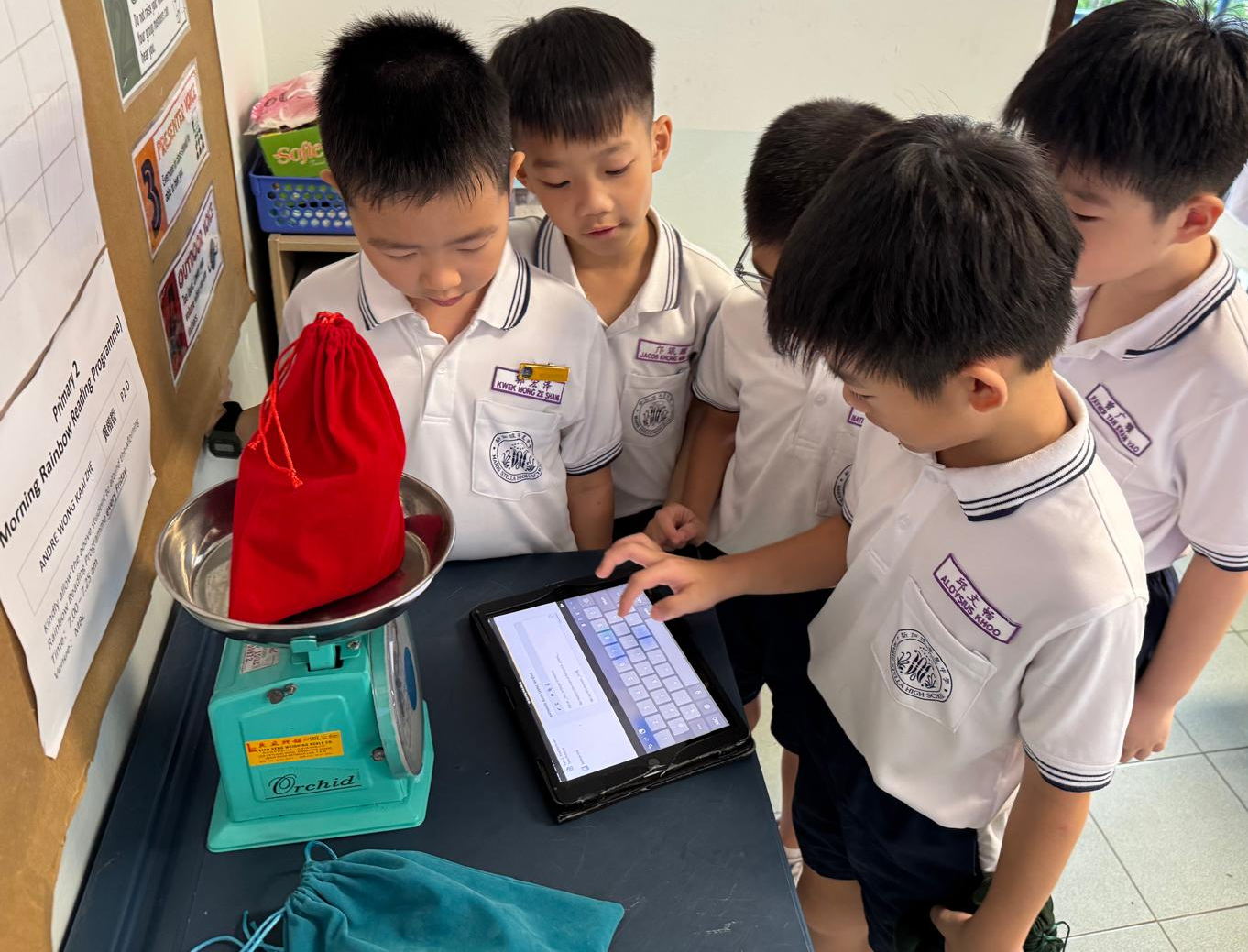 Five young boys carefully weighing a red bag on a small green scale while recording their findings on an iPad.